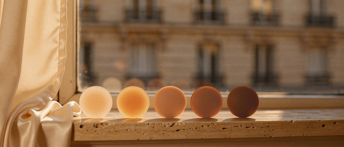 Cinq teintes de cache-tétons en silicone alignées du plus clair au plus profond, Ivoire, Sable, Miel, Noisette, Cacao, sur un rebord de fenêtre parisien à l’heure dorée